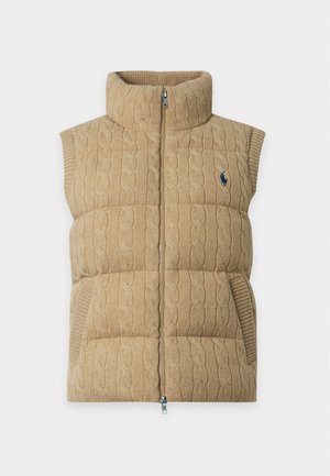 Personne debout avec une main dans la poche, portant un gilet matelassé beige sur un polo en maille blanche à manches courtes et un pantalon large foncé.