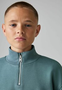 Šviesiai melsvai žalias užtrauktuku užsegamas džemperis su aukšta apykakle, turintis sidabrinį užtrauktuką ir raštuotą tekstūrą. Paprastas ir modernus dizainas.