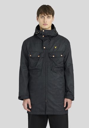 Jeune homme portant une veste noire à capuche avec poches avant et boutons-pression métalliques, debout devant un fond gris clair uni.
