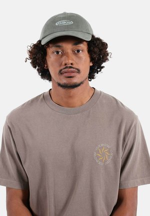 Homme aux cheveux bouclés portant une casquette grise Oxbow et un t-shirt taupe Oxbow avec un logo soleil jaune sur la poitrine, faisant face à la caméra avec un fond uni.