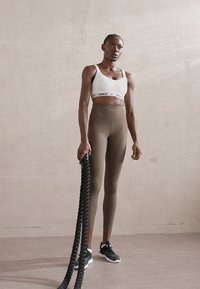 Nike sport brystholder i hvit med logo, brune høytalende leggings og svarte athletic sko. Holder tykke svarte treningstau.
