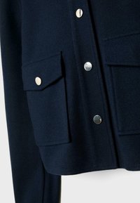 Veste bleu marine foncé avec deux grandes poches à rabat devant et des boutons-pression argentés brillants à l'avant et sur les poches.