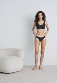 Crni bikini set od rastezljivog materijala; gornji dio s V-izrezom, donji dio s kontrastnim svijetlim akcentima. Manekenka stoji na svijetlom tepihu.