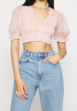 Femme portant un chemisier rose court à manches bouffantes et un jean bleu taille haute, main dans la poche et collier en coquillage.