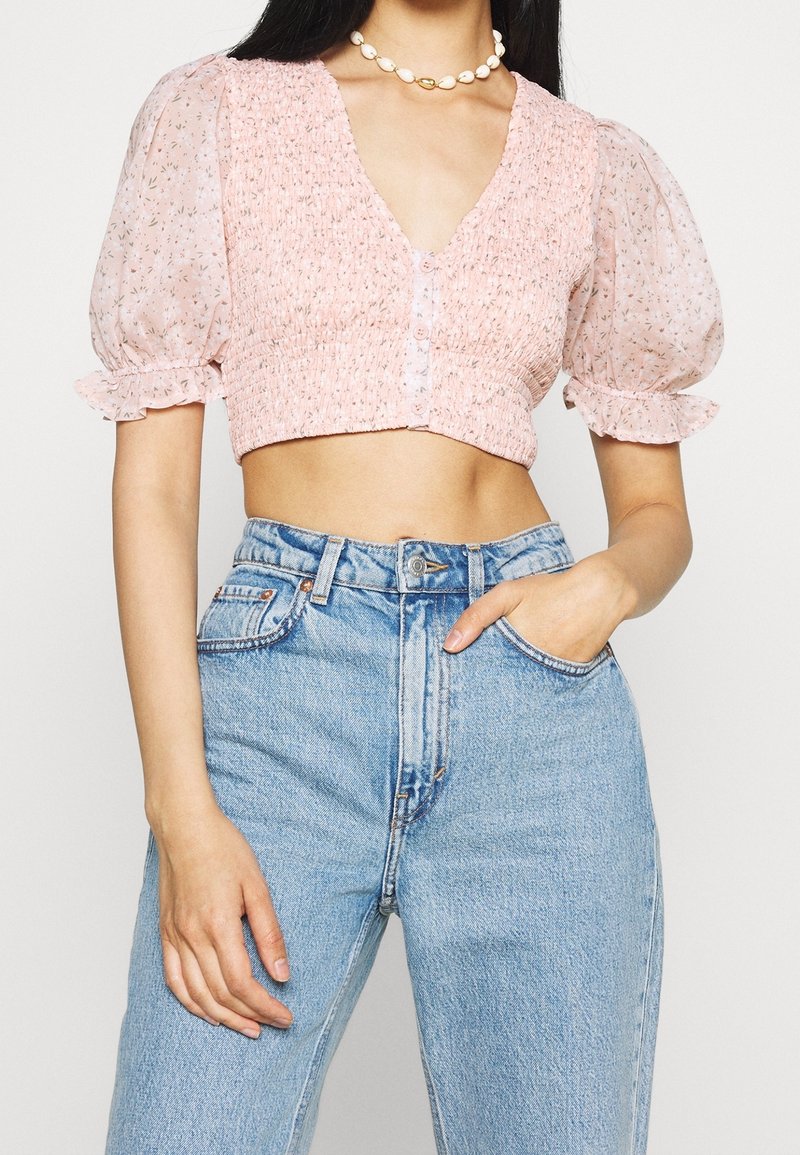 Femme portant un chemisier rose court à manches bouffantes et un jean bleu taille haute, main dans la poche et collier en coquillage.