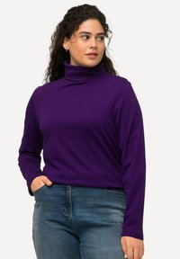 Lila langärmeliger Rollkragenpullover aus einem glatten, dehnbaren Stoff, kombiniert mit blauen Jeans. Das Oberteil hat eine figurbetonte Silhouette und minimale Details.