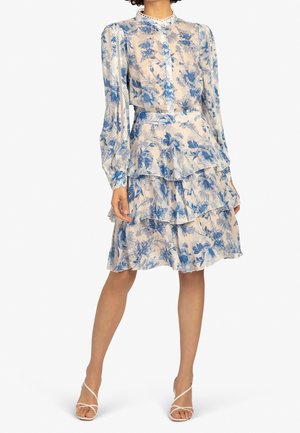 Femme portant une robe fleurie mi-longue à manches longues, avec des volants superposés bleus et blancs, et des sandales blanches à talons hauts et fines brides.