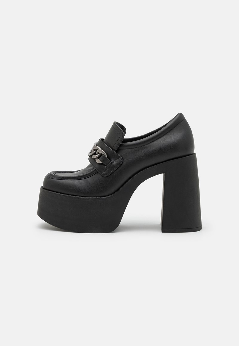 Mocassin à plateforme noire avec un talon haut et épais et une chaîne argentée décorative traversant le dessus, sur un fond clair uni.