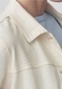 Krémová džínová bunda s překlopeným límcem, viditelným šitím a hladkou texturou, vrstvená přes světle modrý top.