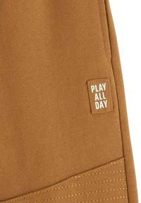 Braune Baumwollshorts mit strukturiertem Design und einem quadratischen Stofflabel, auf dem in kontrastierendem Weiß "PLAY ALL DAY" steht.