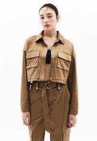 Zkrácená khaki bunda s předním zapínáním na knoflíky, dvěma náprsními kapsami a špičatým límcem. Hladká textura a jemný lesk.