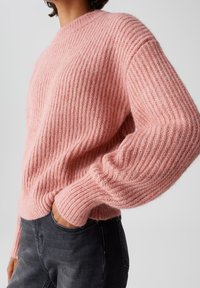 Person, der einen gerippten, rosafarbenen Strickpullover mit langen Ärmeln und schwarze Jeans trägt, mit einer Hand in der Tasche vor einem einfarbigen Hintergrund.