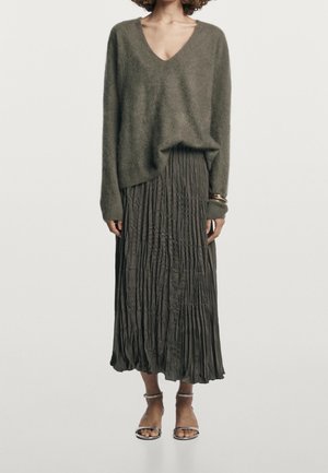 Femme portant un pull doux à col en V vert olive, une jupe midi plissée, des bracelets dorés et des sandales argentées à bout ouvert, se tenant devant un fond blanc.