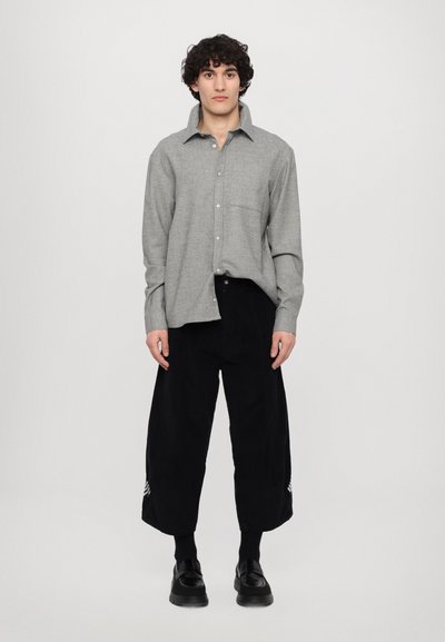 Chemise gris à boutons avec une poche poitrine, associée à un pantalon noir ample avec des bandes sur les côtés, des chaussettes noires et des chaussures noires.