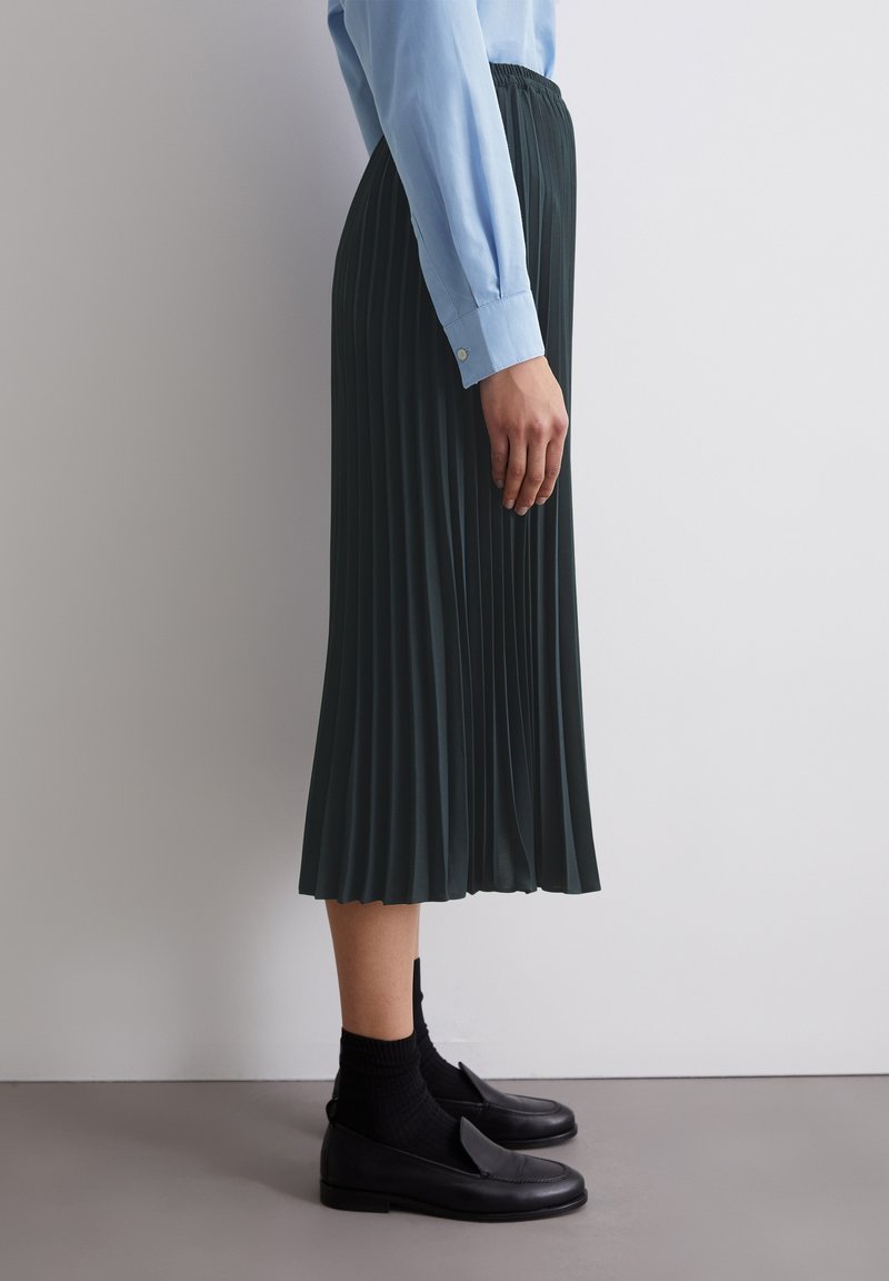 Faltenrock in Dunkelgrün bis zur Mitte mit elastischem Bund, kombiniert mit einem hellblauen Langarmshirt und schwarzen Loafers, seitlich betrachtet.