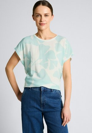 Frau trägt eine hellblaue und weiße kurzärmelige Bluse mit Blumenmuster, die in eine hoch taillierte blaue Denim-Jeans gesteckt ist, steht vor einem schlichten Hintergrund.