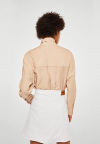 Chemise beige à manches longues avec un détail de empiècement au dos, associée à une jupe en denim blanche avec une étiquette de ceinture et des coutures visibles.