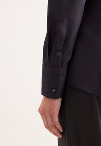 Camicia nera a maniche lunghe con una texture liscia, caratterizzata da un polsino ben rifinito con tre bottoni neri e un design del polsino aperto.