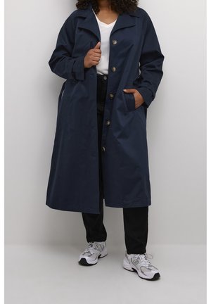 Persoon die een lange marineblauwe jas, wit overhemd, zwarte broek en witte sportschoenen draagt, staand tegen een effen lichte achtergrond.