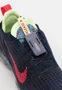 Urheilujalkine, jossa on tummansininen neulepäällinen, punainen Nike-logo, kirkas vihreä sisus ja läpinäkyvä nauhajärjestelmä. Sisältää "RELEASE"-välilehden.