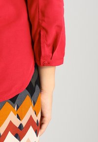 Chemise rouge en coton à manches longues avec poignets boutonnés, associée à une jupe à motifs avec des rayures en zigzag orange, noir et crème.