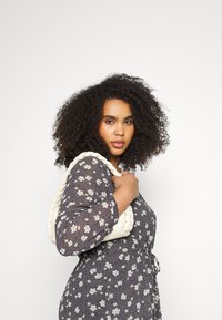 Bolsa trançada branca, feita de material macio, segurada por uma pessoa vestindo um vestido escuro com padrão floral e mangas três quartos, contra um fundo simples.