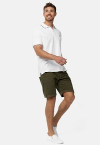 Bela polo majica s kontrastnim ovratnikom, uparen s maslinasto zelenim šortsevima. Model nosi bijele slip-on cipele, stojeći u opuštenom stavu.