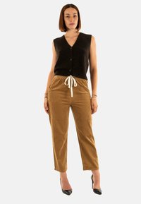 Gilet noir avec boutons associé à un pantalon en velours côtelé marron clair avec cordon de serrage, porté avec des chaussures à talons hauts noires.