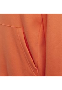 Primo piano del tessuto di una felpa arancione, che mostra una trama liscia, cuciture rifinite e un dettaglio con tasca laterale. Design semplice e minimalista.