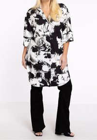 Camisa de gran tamaño en blanco y negro con estampado floral, con botones en la parte frontal y mangas cortas, combinada con pantalones acampanados negros y sandalias de punta abierta.