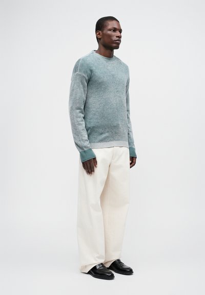 Roheline tekstureeritud kudumine särk ribidega mansettidega, kombineeritud lahtiste kreemivärvi pükstega ja mustade mokkades, seistes neutraalse taustal.