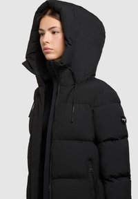 Giacca a puffo nera con colletto alto e grande cappuccio. Presenta una chiusura con zip e tasche laterali. Texture liscia e trapuntata con dettaglio del logo.