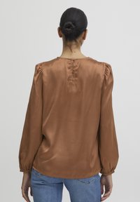 Blusa in satin marrone con maniche a sbuffo, scollatura rotonda e chiusura con bottone sul retro. Tessuto leggero, dalla texture liscia e dalla vestibilità comoda.