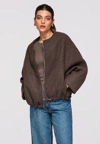 Femme portant une veste zippée marron foncé oversize sur un haut marron et un jean bleu, se tenant les mains dans les poches de la veste devant un fond uni.