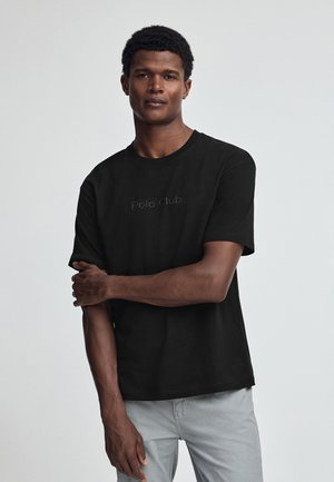 Sort bomuldscrewneck T-shirt med et diskret "Polo Club" logo i midten, korte ærmer og afslappet pasform. Lys grå bukser.
