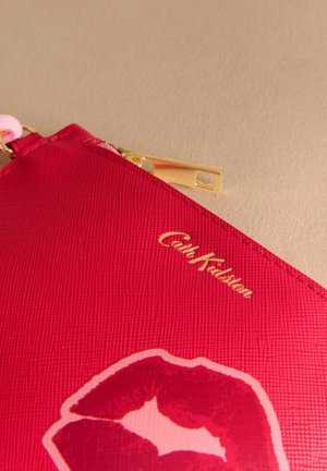 Sarkans teksturēts maisiņš ar zeltījumu apdari, priekšpusē ar lūpu nospieduma dizainu un "Cath Kidston" logotipu zelta burtiem.