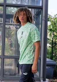 Camiseta verde claro con patrones geométricos, de mangas cortas y cuello redondo. Combinada con pantalones oscuros que muestran un logo.