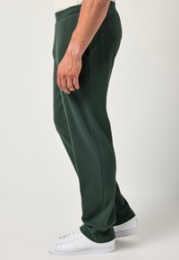 Pantalones de chándal verde oscuro con una textura suave, cinturilla elástica y bolsillos laterales, combinados con zapatillas deportivas blancas.