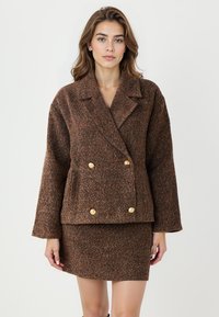 Braune strukturierte Doppelreiherjacke mit großen goldenen Knöpfen, breiten Revers und leicht übergroßen Ärmeln, kombiniert mit einem passenden Rock.