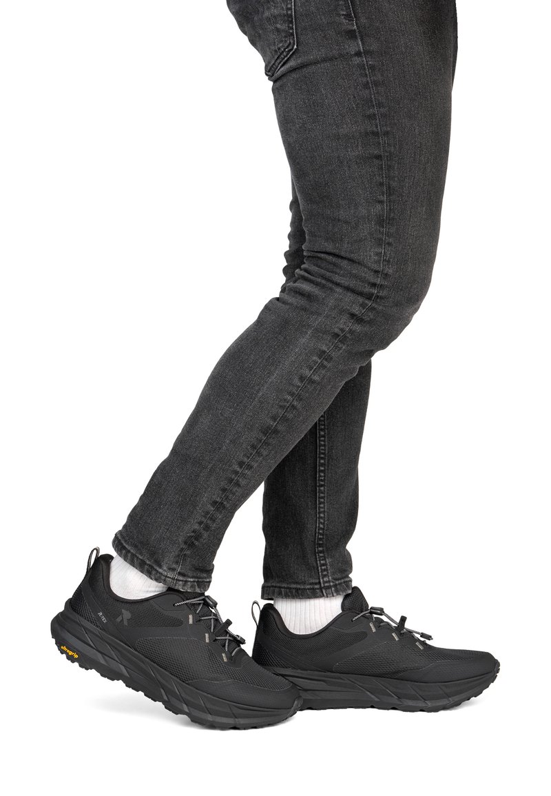 Personne portant des chaussures de sport noires, des chaussettes blanches et un jean skinny gris foncé, debout avec une jambe légèrement pliée.