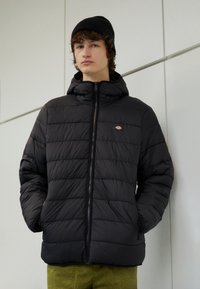 Black puffer jacket with a hood and horizontal stitch pattern; features a front zipper and a small logo on the left chest.