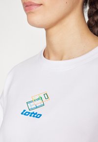 Fehér póló, színes geometriai mintákkal és kék "lotto" felirattal a mellrészen. Az anyaga sima és könnyűnek tűnik.