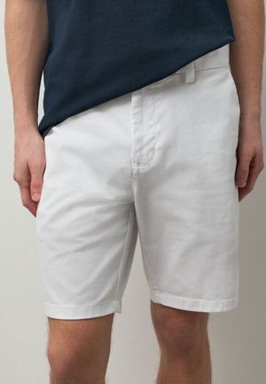 Hombre con pantalones cortos blancos hasta la rodilla y una camiseta azul marino oscuro, de pie con las manos relajadas a los lados.