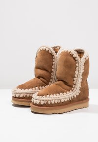Bottes ankle en daim marron avec une doublure en fausse fourrure, bord beige tressé autour du haut et semelle en caoutchouc plate. Texture douce et forme arrondie.