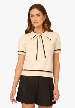Woman wearing a cream short-sleeve top with black trim and bow tie neckline, paired with high-waist black shorts.