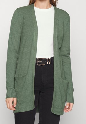 Vrouw draagt een groene open voorkant cardigan over een wit shirt met zwarte broek en een zwarte riem met een metalen gesp.