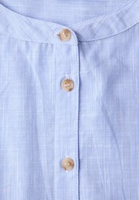 Hellblaues Hemd mit Nadelstreifen, beige Knöpfe und abgerundetem Kragen, zeigt Stoffstruktur und Knopfnaht.