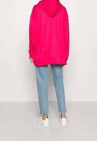 Svetle růžová oversized mikina s kapucí, obsahující klokánkovou kapsu. V kombinaci se světle modrými zúženými džínami a bílými teniskami.