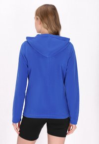 Felpa blu in pile con una texture morbida, maniche lunghe e cappuccio. Indossata sopra dei pantaloni corti neri, caratterizzati da un design semplice e senza loghi visibili.