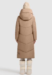 Hosszú, bézs színű puffernadrág kapucnival, célszerű cikkcakk mintával. Krém színű, hajtott mandzsettájú csizmákra viselve.